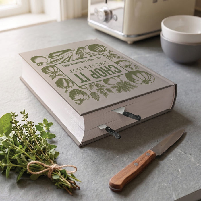 A decorative hardcover book with a green vegetable-themed cover design sits open on a gray countertop, with kitchen knife bookmarks inserted in the pages. A small bunch of fresh herbs and a wooden-handled knife are placed nearby, with a cream appliance and stacked bowls in the softly blurred background.

Cookmarks - mini chef knife bookmarks - make a great gift under $10 for home chefs, cooks, and cookbook lovers.