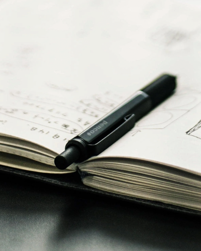 A black Pilot ballpoint pen resting on an open notebook with handwritten and sketched pages.