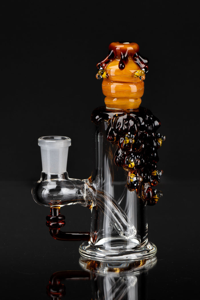 Close-up, three-quarter view of the Mini Rig - Beehive Nano perched atop a clear glass rig with amber honeycomb and glossy drips against a black background