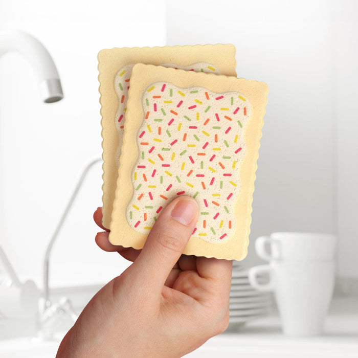 A hand holding Fred Slop Tarts Compressed Kitchen Sponges in front of a kitchen sink