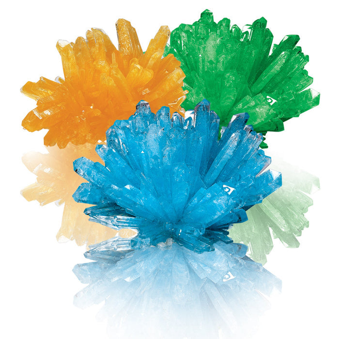 Three vibrant crystal clusters—orange, green, and blue—grown with the Smithsonian Crystal Growing kit by Smithsonian, are grouped on a white background with reflections beneath them. Ideal for a fun STEM project.